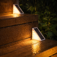 Two triangular solar outdoor lights glowing warmly on wet wooden steps in the rain, with blurred green foliage in the background.