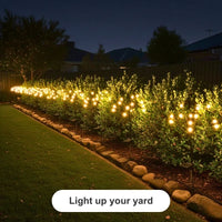 Outdoor garden scene at night with many small warm white globe lights attached to bushes along a stone-edged lawn border. A dark fence and houses are visible in the background, and a white label at the bottom reads 'Light up your yard'.